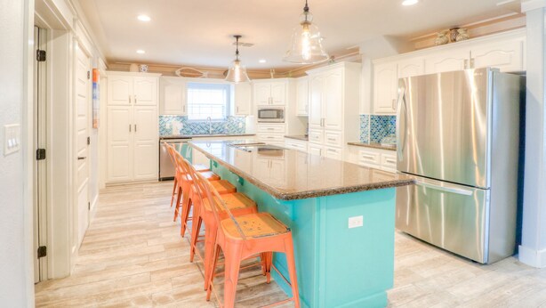 A modern kitchen with white cabinets, a central island with bar stools, a stainless steel refrigerator, and a tiled backsplash. - Stay 7+ Nights for 10% Off: Isla Breeze, Your Easeful Escape (South Padre Island)