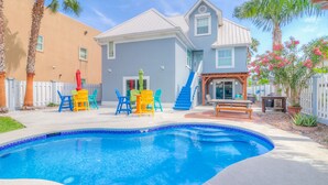 A swimming pool with a blue tiled edge, a patio area with colorful chairs, and a house with a light blue exterior and a white fence. - Stay 7+ Nights for 10% Off: Isla Breeze, Your Easeful Escape (South Padre Island)