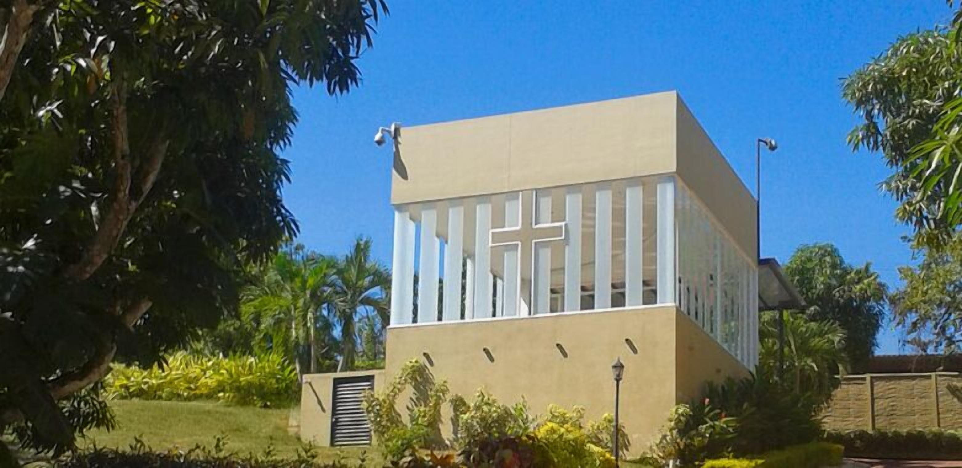 Un bâtiment moderne orné d’une croix sur sa façade, entouré d’une végétation luxuriante.