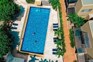 Eine Vogelperspektive zeigt einen rechteckigen Swimmingpool, umgeben von Palmen und Liegestühlen, mit einer gefliesten Terrasse und einem an das Becken angrenzenden Gebäude.
