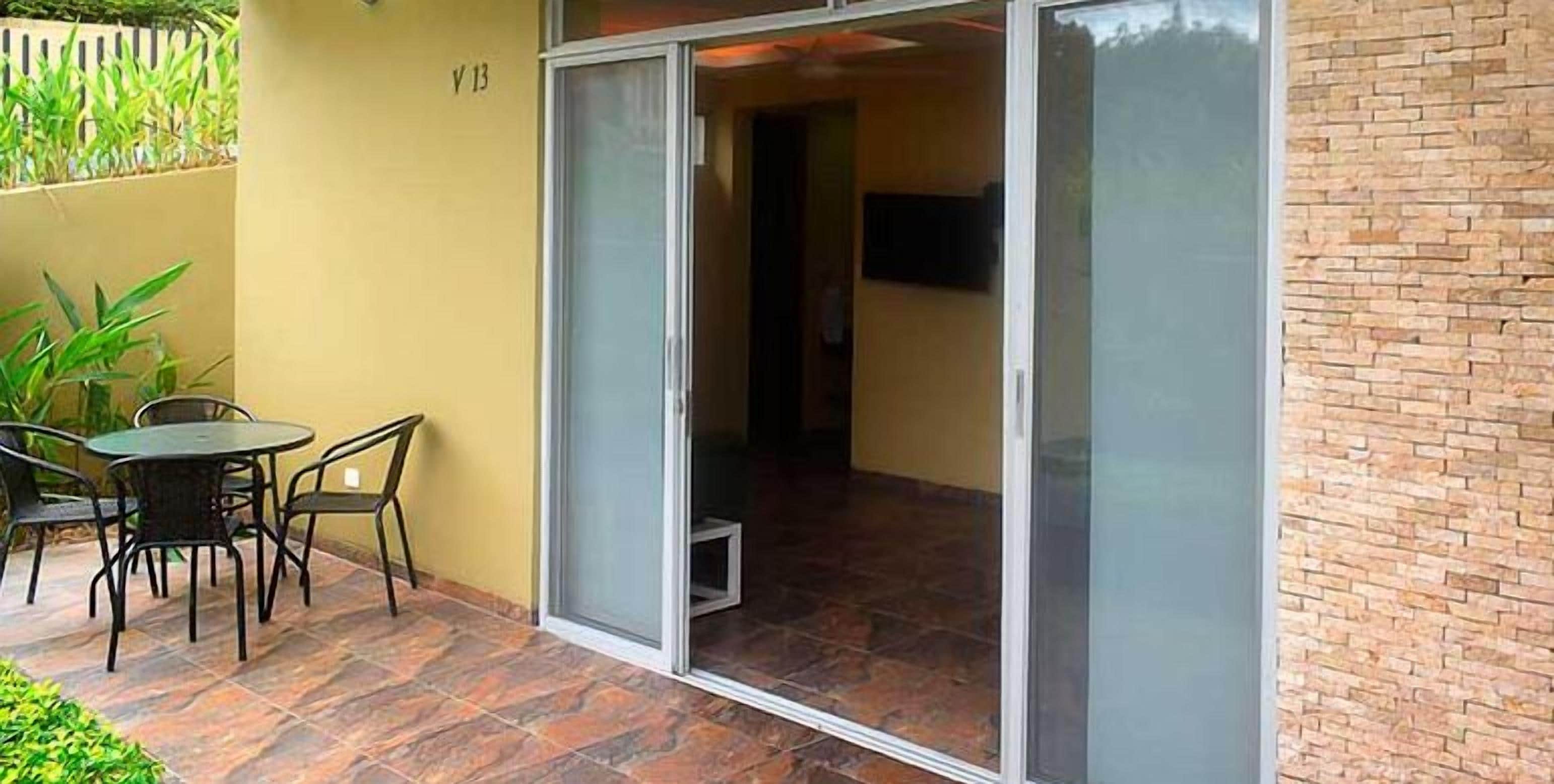 Een patio met een tafel en stoelen, een schuifdeur naar een binnenruimte, en een bakstenen muur aan de rechterkant.