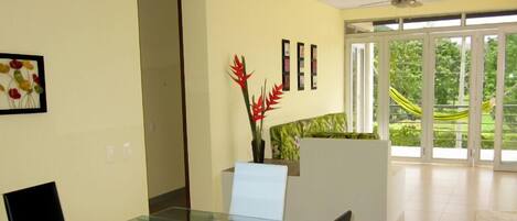 A modern interior with a glass dining table, a ceiling fan, and a view of greenery through large sliding doors.