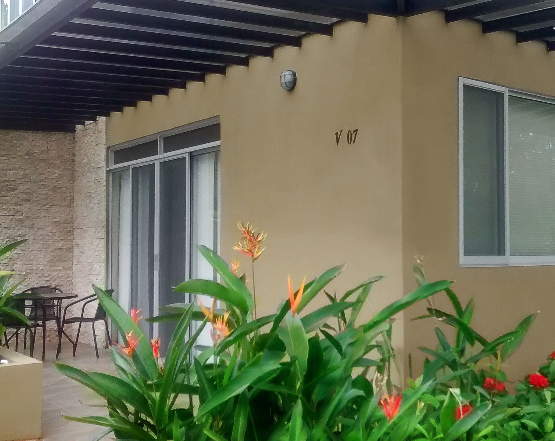 A covered outdoor area with a beige wall, a window, and a door. There are plants and flowers in the foreground.