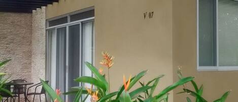 A covered outdoor area with a beige wall, a window, and a door. There are plants and flowers in the foreground.