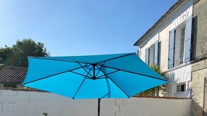 Una terraza de madera con dos sillones de descanso y una mesa, una planta en maceta y un amplio paraguas azul que proporciona sombra.