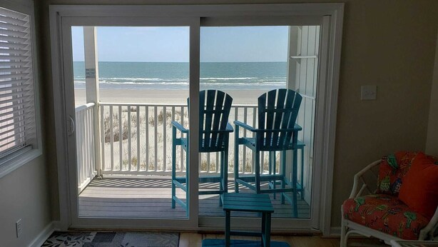 Een kamer met uitzicht op het strand, een schuifdeur, twee blauwe Adirondack-stoelen en een rode stoel met een kleurrijk kussen.