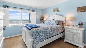 Een slaapkamer met een bed, nachtkastjes, een plafondventilator, een raam met uitzicht op het strand en een inspirerende muurposter.