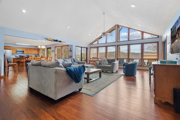 A spacious living area with a large window, a kitchen in the background, and a ceiling fan.
