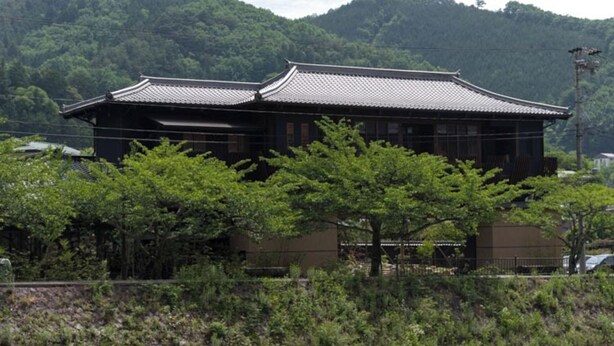 A traditional Japanese building with a tiled roof, surrounded by greenery and hills. - Kurotake (Achi)