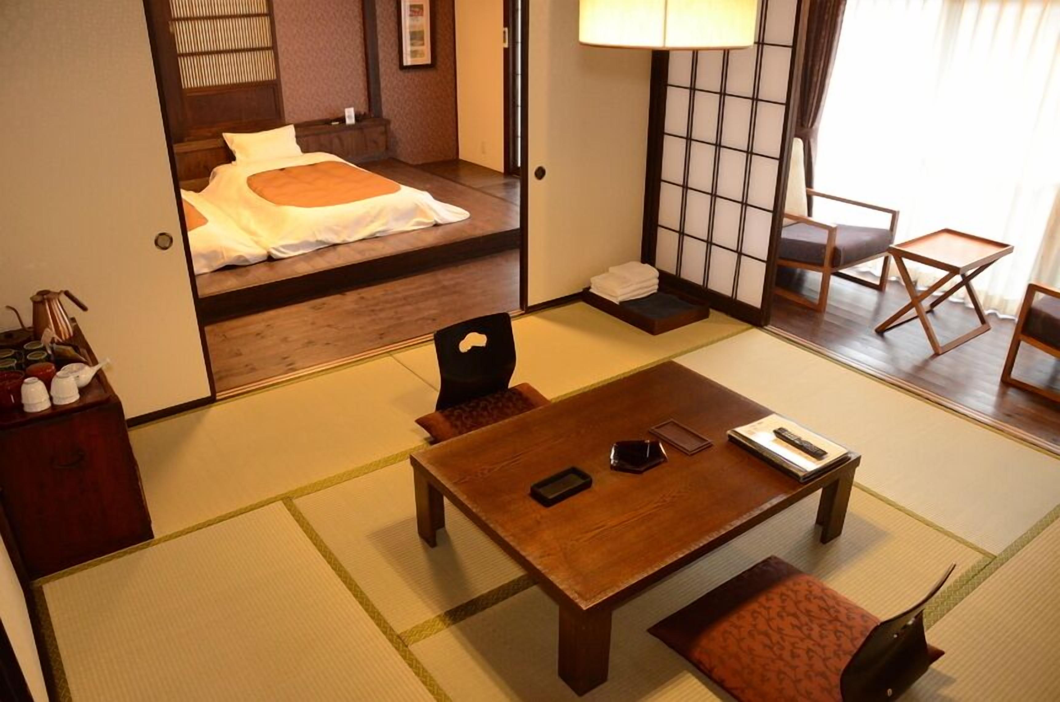 A traditional Japanese room with tatami flooring, a low table with cushions, a bed with a white and orange bedding, and a sliding door leading to another room.