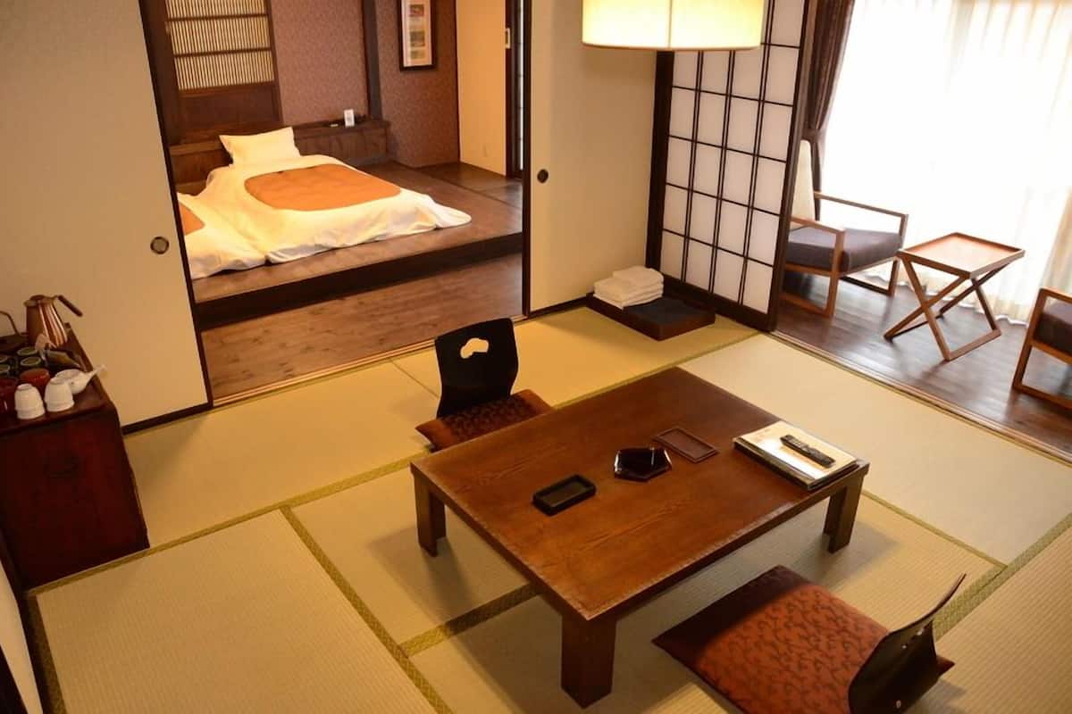 Une pièce de style japonais traditionnel, avec un sol recouvert de tatamis, une table basse avec des coussins, un lit avec une literie blanche et orange, et une porte coulissante donnant sur une autre pièce.