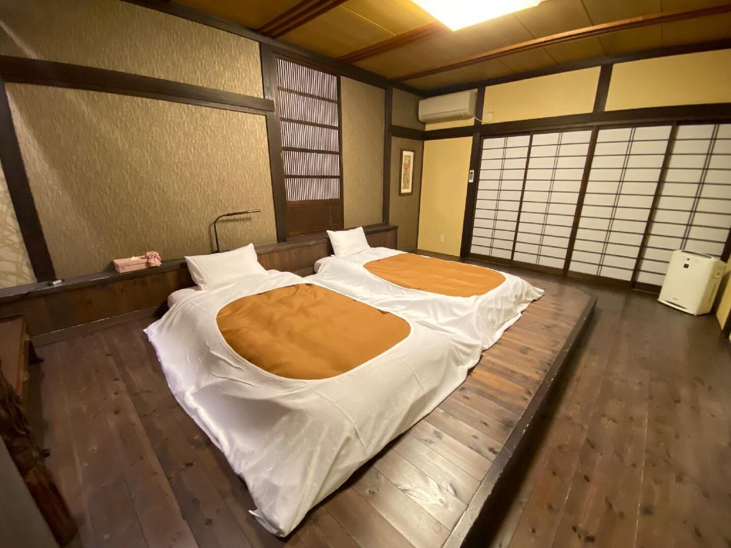 A traditional Japanese-style room with tatami flooring, two beds with white bedding and brown accents, a shoji screen, and a small air conditioning unit.
