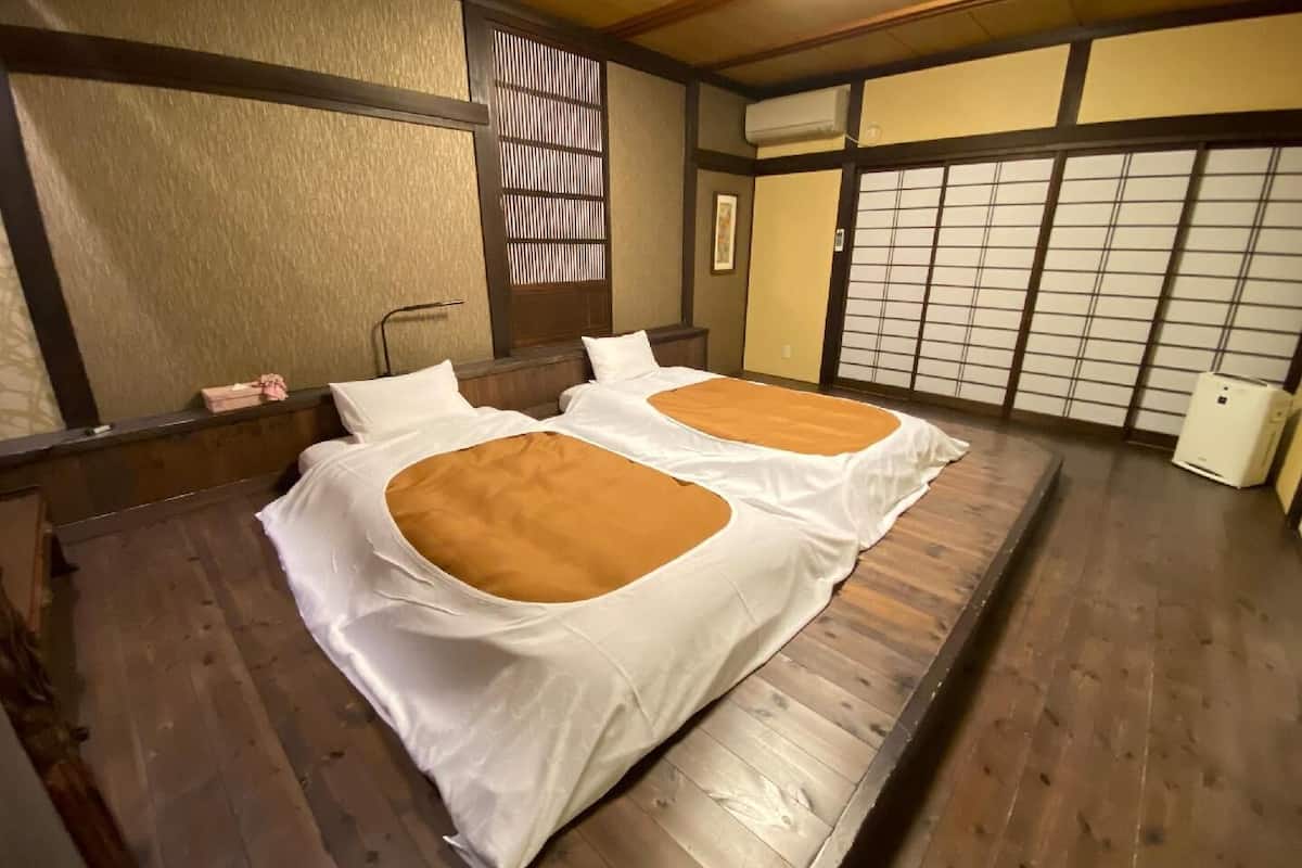 Une pièce de style japonais traditionnel, avec un sol recouvert de tatamis, deux lits avec une literie blanche et des touches de couleur marron, un paravent shoji et un petit climatiseur.