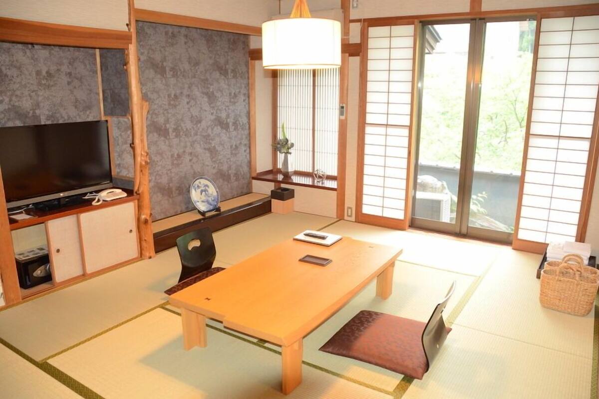 A traditional Japanese room with tatami flooring, a low table, a television, sliding doors, and a view of greenery outside.