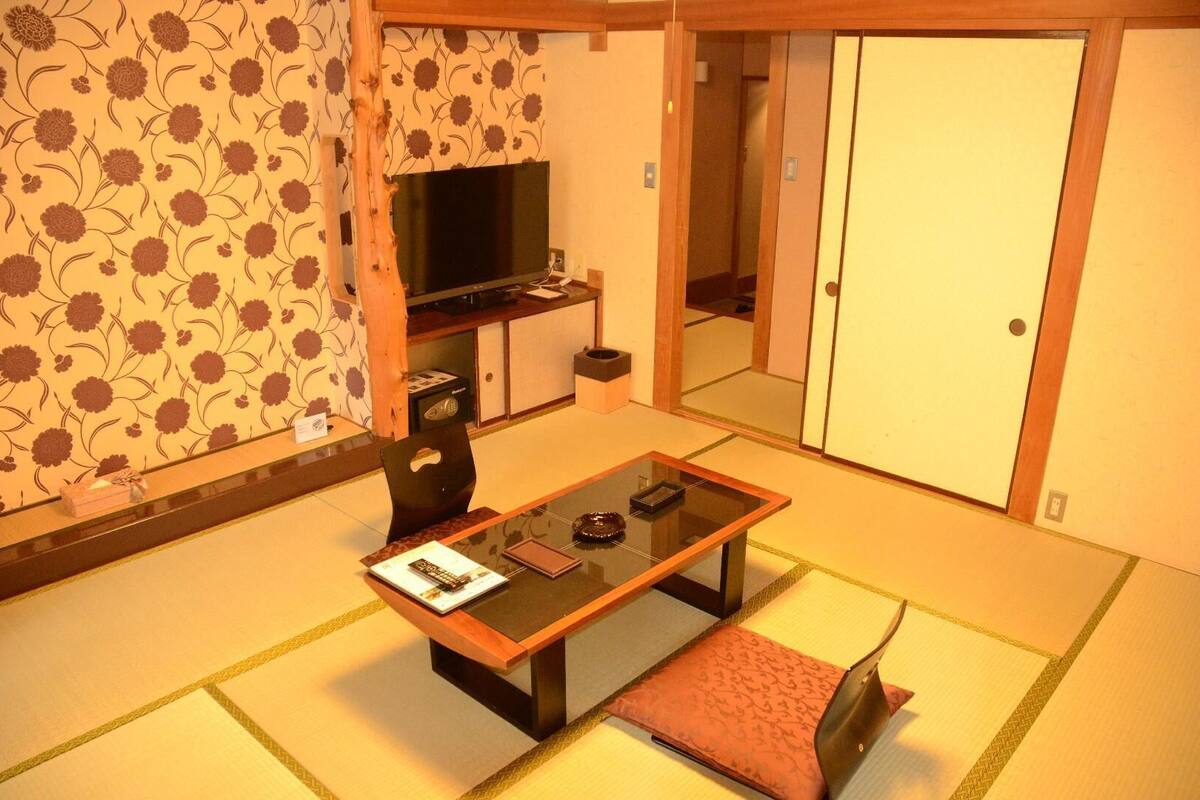 Une pièce de style japonais traditionnel, avec un sol recouvert de tatamis, une table basse avec des coussins, un téléviseur et des portes coulissantes.