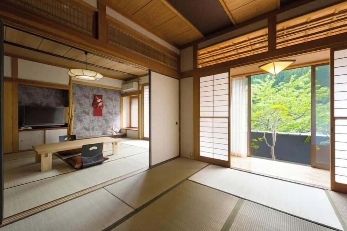 A traditional Japanese room with tatami flooring, a low table, a television, and sliding doors with a view of greenery.