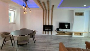 Een moderne woonkamer met een ronde eettafel, zachte zitplaatsen, een flatscreen televisie en een plafond met zichtbare balken en inbouwverlichting.