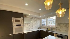 Een moderne keuken met een wit aanrecht, donkere kastjes, een magnetron en een dubbele oven. Boven de spoelbak hangen hanglampen en een raam zorgt voor natuurlijk licht.