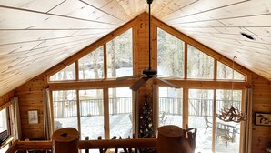 Une cabane dotée d’un plafond en bois incliné, de grandes fenêtres, d’un ventilateur de plafond et d’un coin repas avec des sièges à l’extérieur.