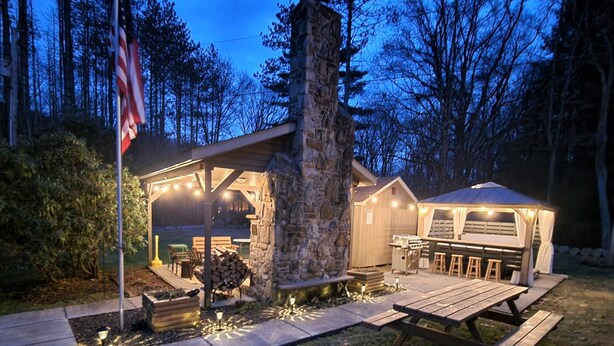 A rustic outdoor patio area with a stone chimney, wooden benches, and a covered barbecue station. - Play & Stay near State Parks & Seven Springs (Champion)