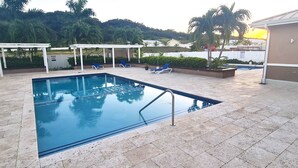 Et rektangulært pool med flisebelagt solterrasse, omgivet af liggestole og et overdækket siddeområde.