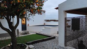 Un área de piscina moderna al aire libre con un pequeño jardín, un árbol de naranjos y un edificio blanco con una puerta negra.