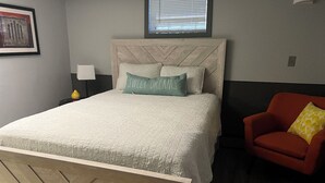 Ein Schlafzimmer mit einem Bett, einem roten Sessel, einem Nachttisch, einer Lampe und einem Fenster mit Jalousien.