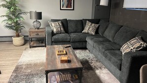Ein modernes Wohnzimmer mit einem Ecksofa, einem Holztisch, einem Beistelltisch, einer Lampe und einem gerahmten Bild an der Wand.