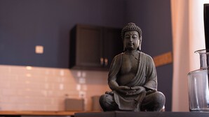 Eine Statue einer sitzenden Figur in einem Raum mit dunklen Wänden, einer gefliesten Rückwand und einer hängenden Leuchte.