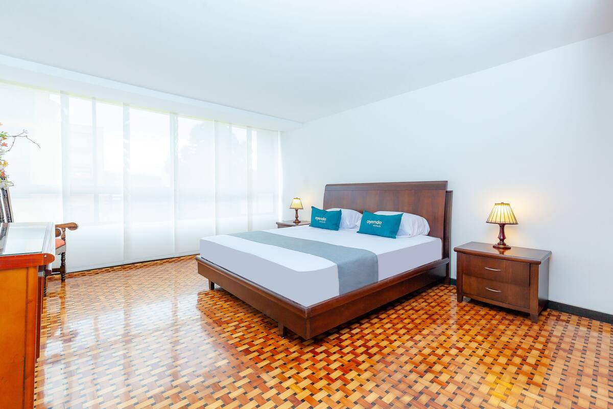 Una habitación de hotel moderna con suelo de parquet de madera, una cama grande con ropa de cama azul y blanca, una mesita de noche con lámpara y un ventanal que proporciona luz natural.