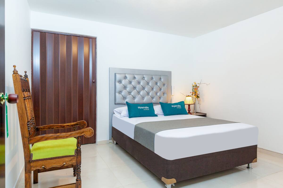 Una habitación de hotel con una cama grande, una silla de madera, un cabecero tapizado y una mesita de noche con lámpara.