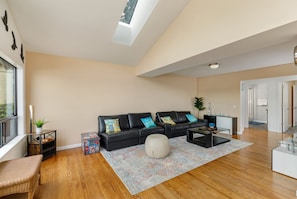 Uma sala de estar ampla com um sofá de couro preto, um banquinho branco e uma mesa de centro. Há uma janela grande que oferece luz natural e vista para o exterior.