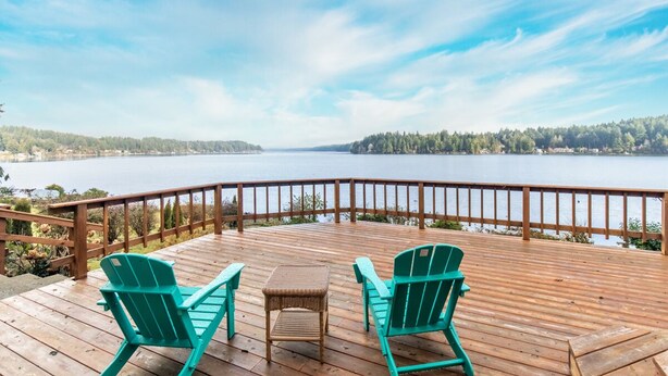 Eine Holzterrasse mit zwei grünen Adirondack-Stühlen und einem kleinen Tisch, mit Blick auf einen ruhigen See mit bewaldeter Uferlinie.