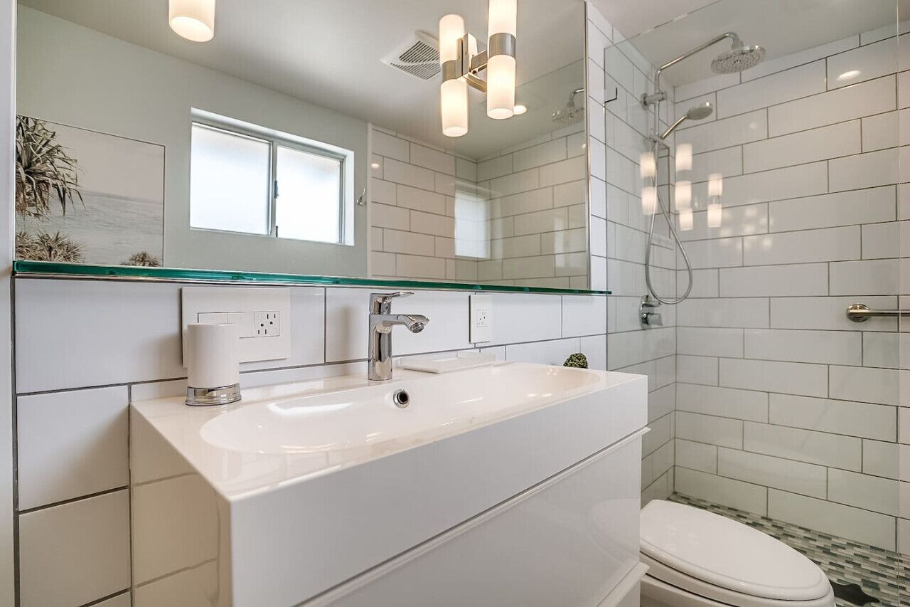 A modern bathroom with a large white sink, a glass countertop, a mirror above the sink, a shower with a handheld showerhead, and a toilet.