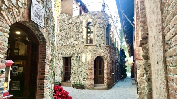 Une ruelle étroite pavée de pierres, bordée de bâtiments en pierre, avec une enseigne indiquant un établissement de jeux et une porte sur laquelle est apposée une autre enseigne.