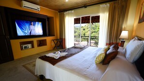 Ein Hotelzimmer mit Bett, Fernseher, einem Balkon mit Vorhängen und Blick auf Grünflächen.
