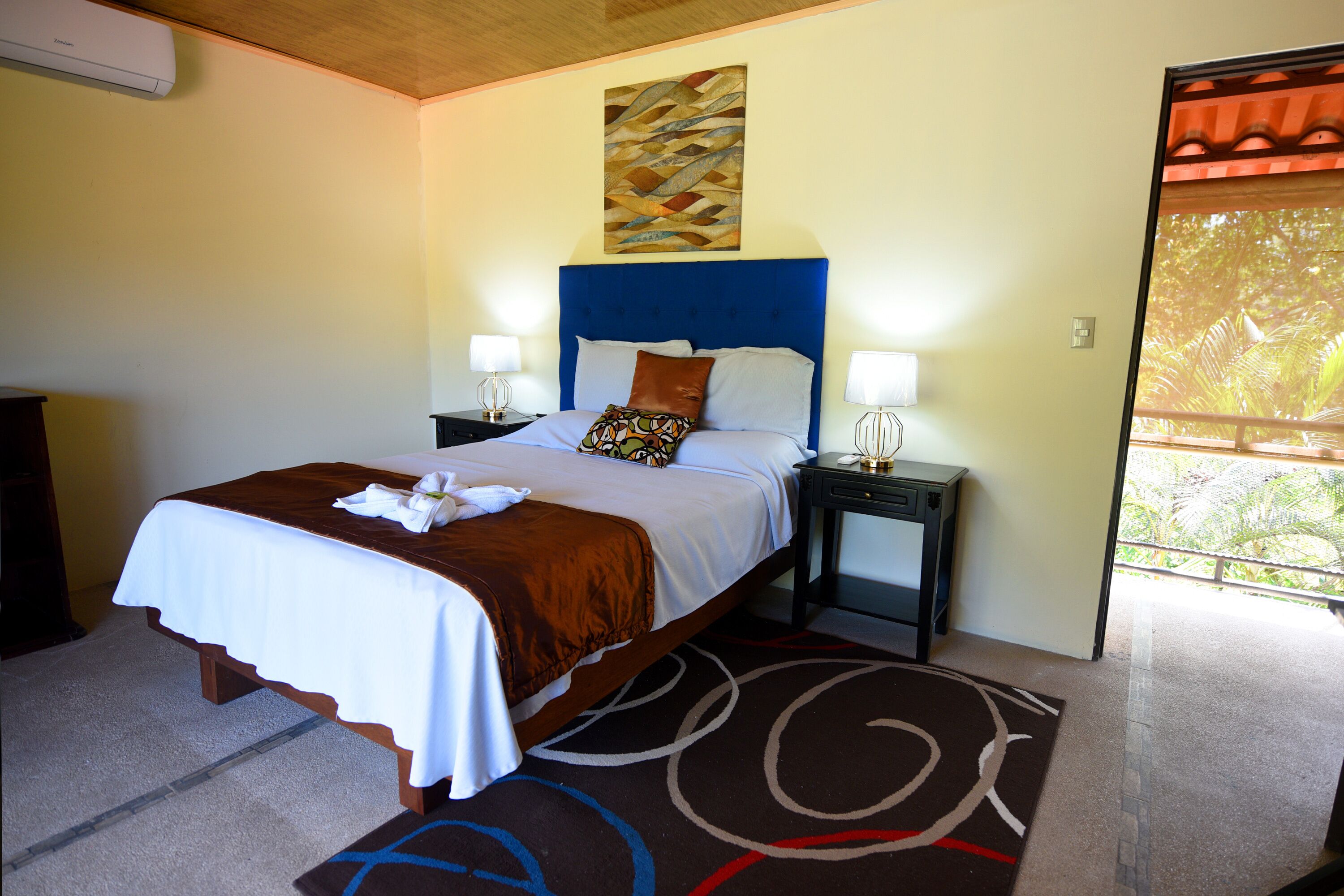 A neatly made bed with a white and brown bedspread, two bedside lamps, a decorative wall piece, and a view of greenery through a window.