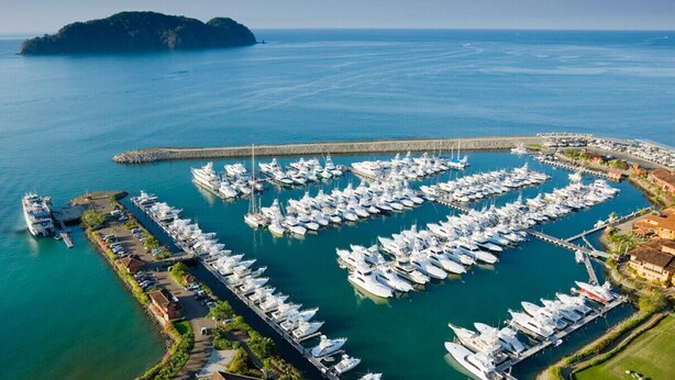Eine Luftaufnahme eines Yachthafens mit zahlreichen vertäuten Booten, umgeben von einer Küstenstadt mit Gebäuden und einem Steg, der sich ins Wasser erstreckt.
