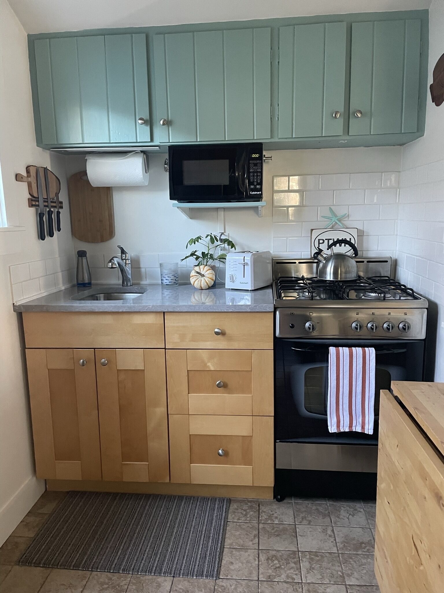 Cocina compacta con gabinetes verde azulado, un microondas, un tostador, un hervidor y una estufa a gas.