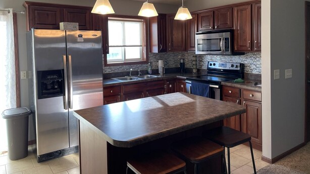 A modern kitchen with stainless steel appliances, wooden cabinets, a large island with bar stools, and pendant lighting. - Cozy ranch style house in a quiet neighborhood (Sioux Falls)