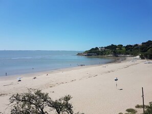 Une vaste plage de sable fin bordée d’une eau d’un bleu limpide, quelques bateaux et quelques personnes. On aperçoit des arbres et des bâtiments au loin.