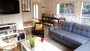 A living room with a grey sofa, a white coffee table with a plant, a TV, and a dining area with a table and chairs.