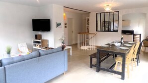 A modern living room with a sofa, TV, and entertainment unit. A dining area with a table and chairs is visible.