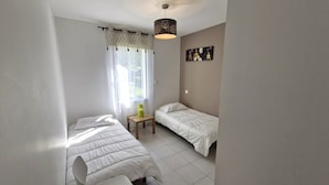 Una habitación con dos camas individuales, una mesita pequeña, una silla, una lámpara colgante y una ventana con cortinas.