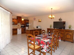 Un'area pranzo con tavolo imbandito per quattro persone, sedie di legno, un lampadario, un televisore e una cucina sullo sfondo.