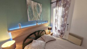 Ein Schlafzimmer mit einem Holz-Kopfteil, einem Bett mit weißer Bettwäsche, einem Nachttisch mit Lampe, einem Kaktus auf dem Fensterbrett und einem großen Fenster mit Vorhängen.