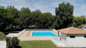 Ein rechteckiger Pool mit Fliesen umgeben, von einem niedrigen Zaun begrenzt, vor einer dichten Baumlandschaft und blauem Himmel.