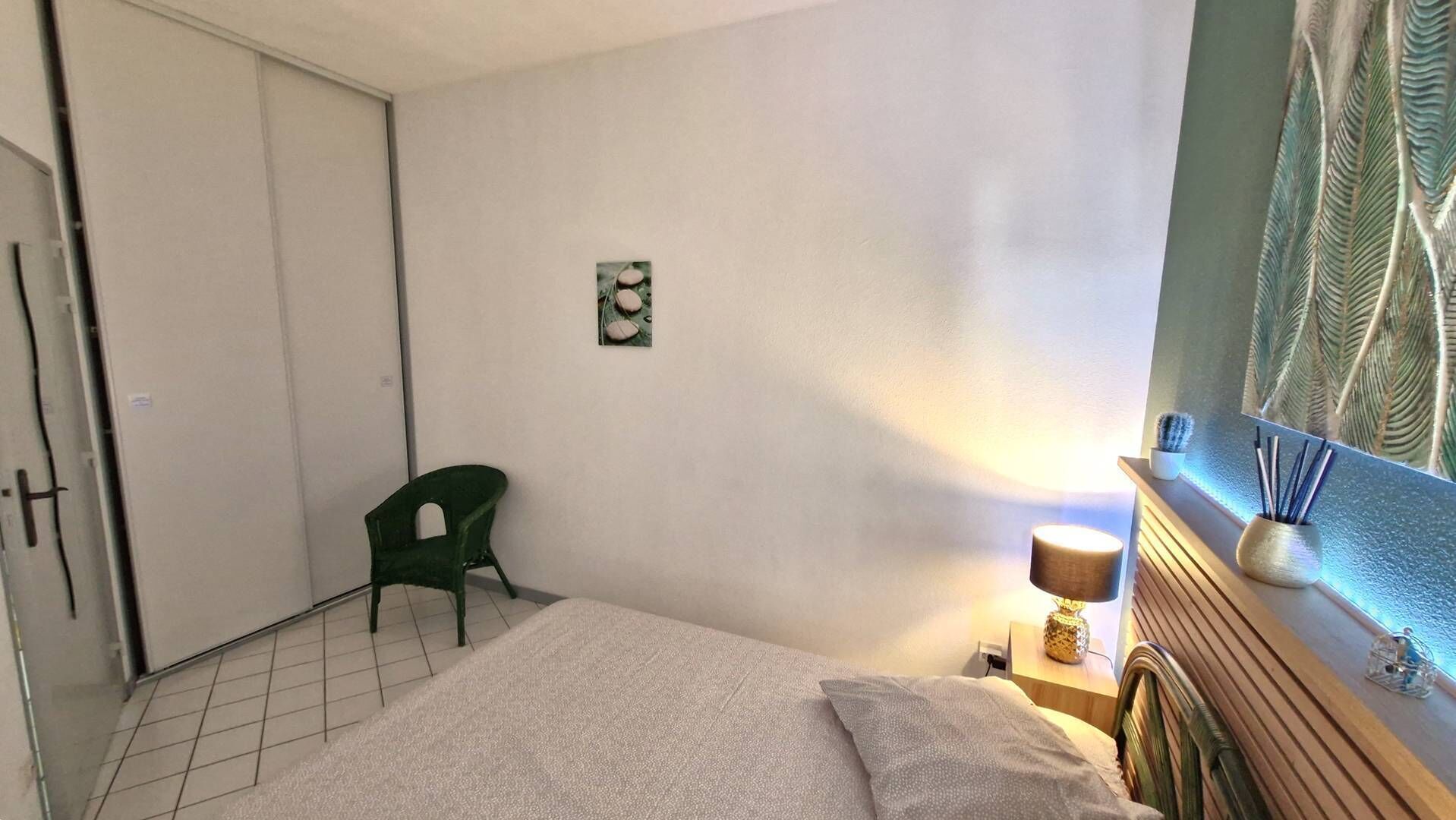 A bedroom with a bed, a green chair, a small table with a lamp, and a wall-mounted shelf with decorative items.