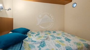 A bedroom with a bed, two blue pillows, a floral-patterned bedspread, wooden headboard, and a wall-mounted light fixture. - AS21387-Studio cabin and private parking 200m sea (LE GRAU DU ROI)