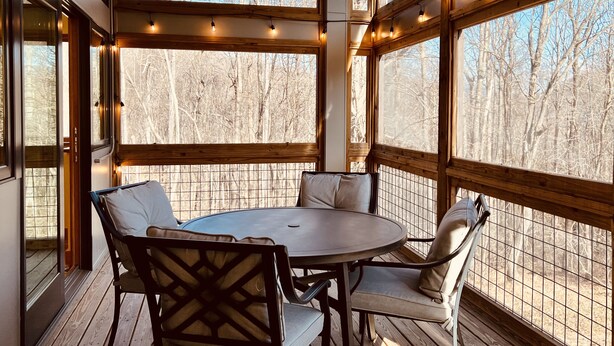 A covered outdoor seating area with a round table and chairs, large windows, and string lights.